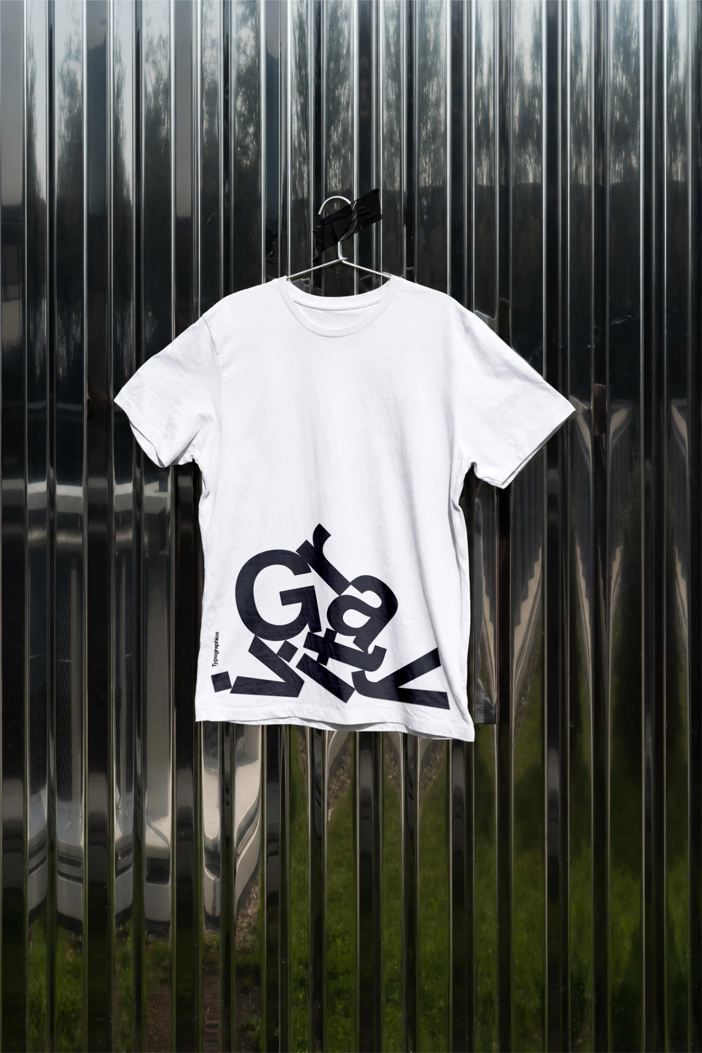White Gravity T-shirt with the small vertical Typographico logo, hanging against a reflective metal wall.