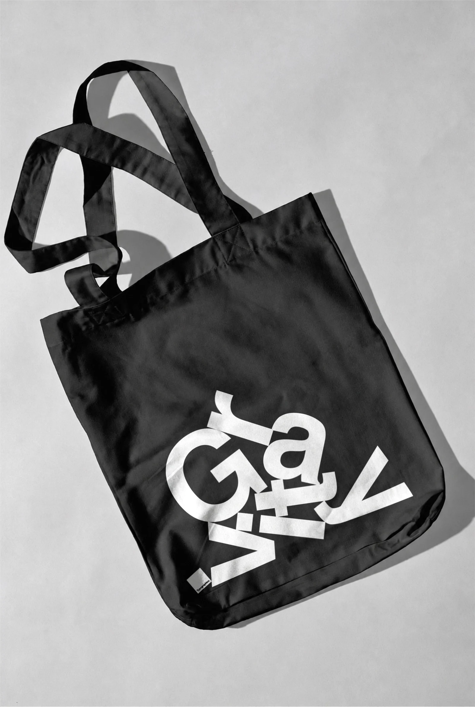 Black tote bag with a white Gravity print, lying on a light grey surface.