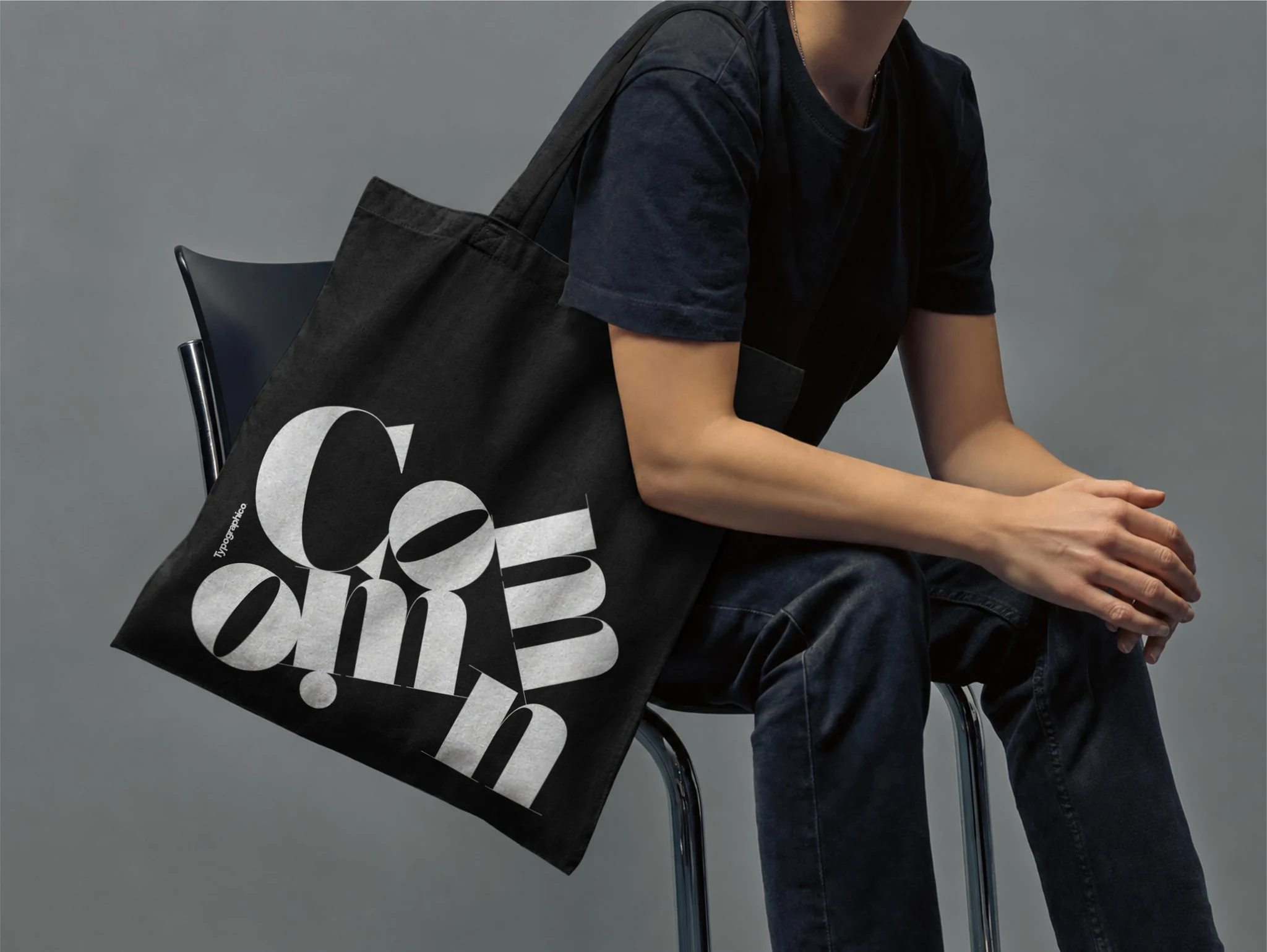 Black Common tote worn over the shoulder by a seated man against a grey background.